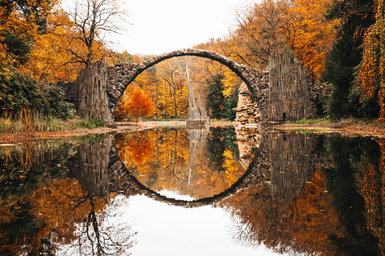 View of The Devil's Bridge reflecting in the calm water of the Rakotzsee surrounded by vibrant autumn foliage in Gablenz, Sachsen, Germany.