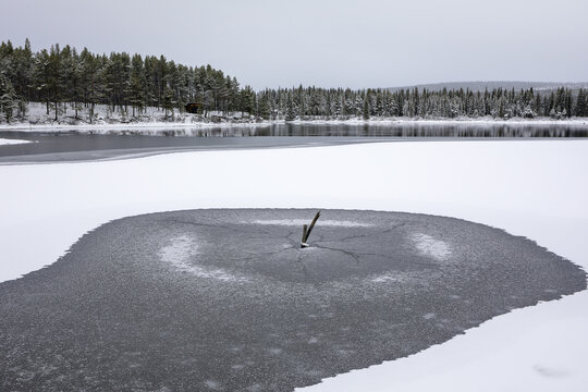 View of a frozen lake with a circular patch of thin ice in the foreground, surrounded by snow-covered ground and a pine forest in Abisko, Norrbotten County, Sweden.