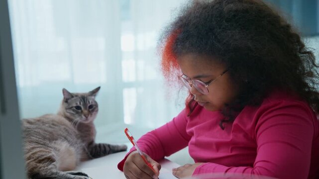 Young girl studies with cat nearby. Girl wears glasses and pink shirt. Cat rests calmly beside her. Focused, quiet, and peaceful. Ideal for education, home, or calm work scenes