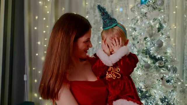 Warm, joyful Christmas moment captured indoors. Young child playfully covers eyes while being held. Mother smiles gently, enjoying festive holiday. Bright, decorated tree glows softly in background