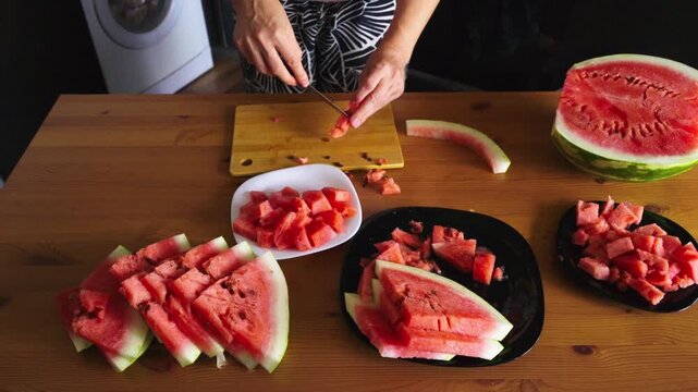 Woman chops watermelon on wooden table. Fresh red fruit cuts in pieces with sharp knife. Plates filled with juicy chunks of watermelon. Simple, bright kitchen scene for summer or healthy eating