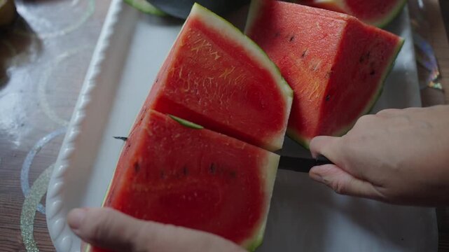 Hands cut watermelon slices on a white tray. Fingers hold fruit, knife cuts through red flesh. Fresh red flesh shows black seeds inside. Summer meal preparation scene with simple focus
