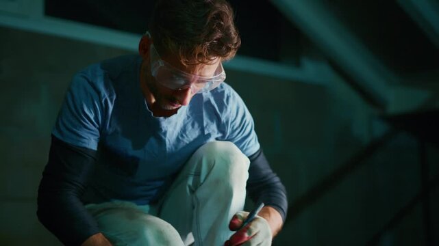 Man wears safety goggles and gloves. He concentrates on marking metal with a tool. Dark industrial space surrounds him. Focused worker prepares metal component for fabrication. Ideal for manufacturing