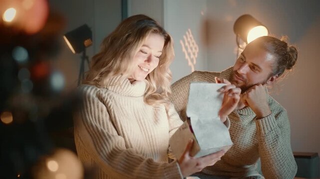 Cozy living room, warm lighting. Couple shares joyful gift moment, smiles brightly. Soft knitted sweaters, intimate, festive setting. Perfect for holiday, romance, or celebration projects