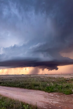 View of a dramatic supercell storm with dark rotating clouds and heavy rain over a vast open field at sunset.