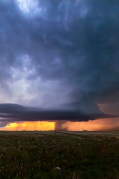 View of a powerful supercell storm with a funnel cloud over a grassy plain under dark, swirling clouds with a golden sunset glow World.