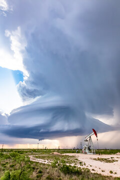 View of a dramatic supercell storm with a rotating cloud formation looming over an oil pump jack in a vast, arid landscape.