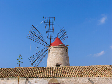 View of the historic saltworks windmill with its distinctive red roof and lattice blades, set against a clear blue sky in Trapani, Sicily, Italy.