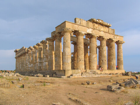 View of the ancient Greek Temple E at Selinunte Archaeological Park with weathered stone columns and people sitting on the steps under a clear blue sky in Marinella, Sicilia, Italy.