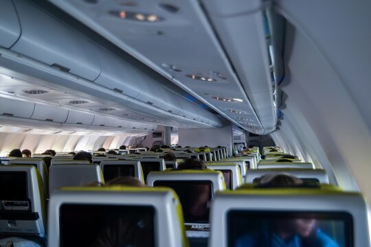 View of airplane cabin interior with rows of passenger seats, overhead storage bins, and seat-back entertainment screens in an aircraft flying over the Atlantic Ocean.