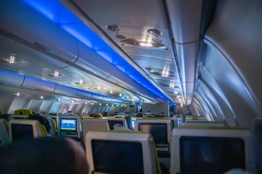 View of an airplane cabin interior with rows of passenger seats, seatback screens, and blue ambient lighting over the Atlantic Ocean.