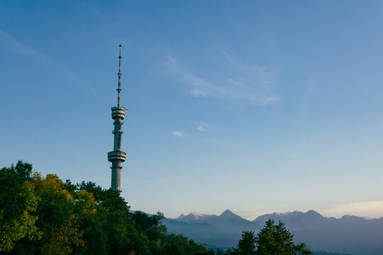 View of the Almaty TV Tower rising above lush green trees with snow-capped mountains in the background in Almaty, Almaty Region, Kazakhstan.