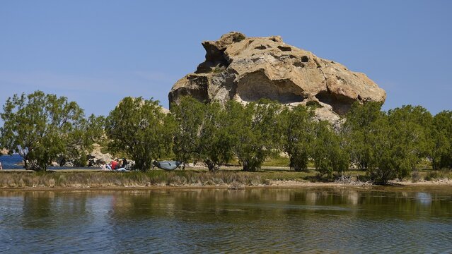 Rock rises behind a row of trees on the lakeshore under a clear sky, Rock of Kalikatsou, Seasonal Salt Lake, Patmos, Dodecanese, Greek Islands, Greece