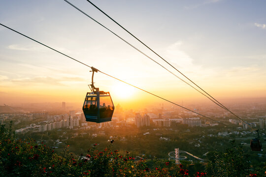 View of a cable car gondola gliding over the city during a golden sunset with mountains in the distance and flowers in the foreground in Almaty, Almaty Region, Kazakhstan.