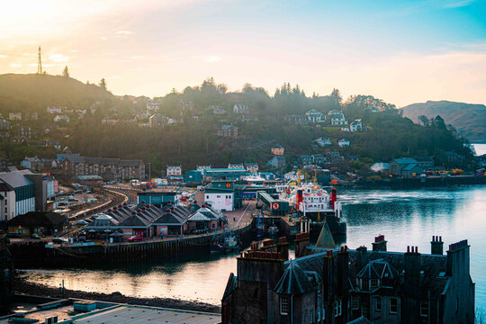View of the harbour with ferry boats docked at the pier, historic stone buildings along the waterfront, and hills in the background in Oban, Scotland, United Kingdom.