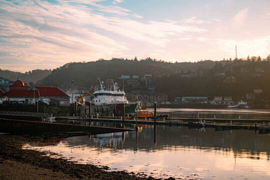 View of the harbor with fishing vessels docked at the pier, historic buildings along the waterfront, and calm water reflecting the soft light in Oban, Scotland, United Kingdom.
