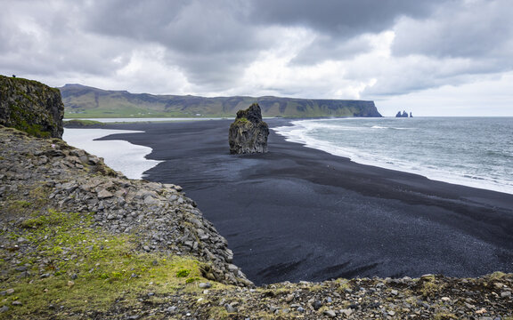 View of Dyrholaey and the dramatic black sand beach with dark volcanic sand, crashing waves, and rugged cliffs under a cloudy sky in Black Sand Beach, Myrdalshreppur, Iceland.