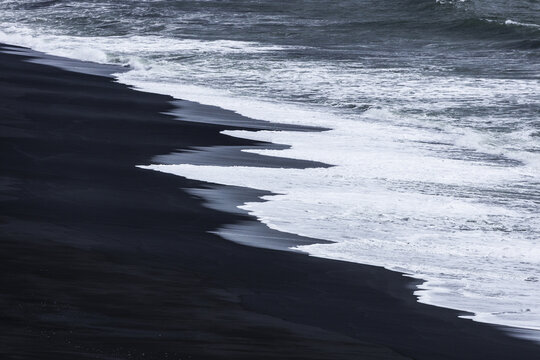 View of the dark volcanic sand and white sea foam washing onto the shore near Dyrholaey, Black Sand Beach, Myrdalshreppur, Iceland.