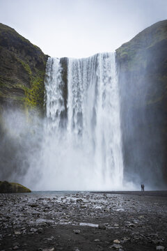 View of Skogafoss waterfall cascading down mossy cliffs with two people standing on the rocky shore in Skogafoss, Rangarthing eystra, Iceland.