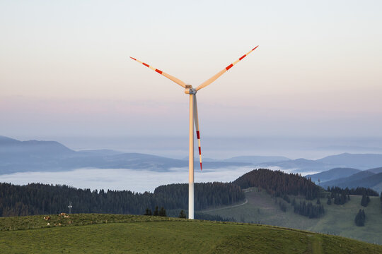View of the Teichalm wind turbine standing on a grassy hill above a forest with a sea of clouds in the background at dawn in Plankogel, Styria, Austria.
