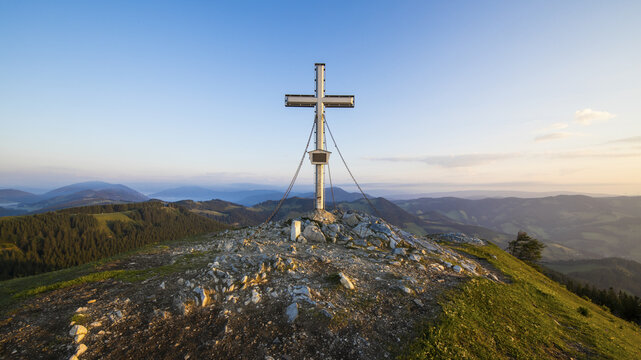 View of the summit cross on Plankogel near Teichalm, featuring rocky terrain, green mountain slopes, and rolling hills under a clear golden hour sky in Plankogel, Styria, Austria.