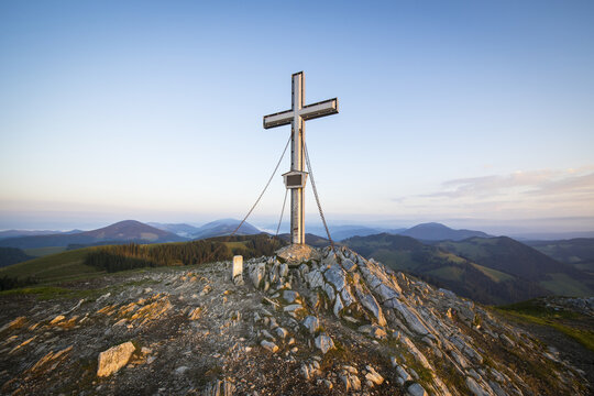 View of the summit cross on Plankogel surrounded by rocky terrain and rolling mountain landscape at golden hour in Plankogel, Styria, Austria.