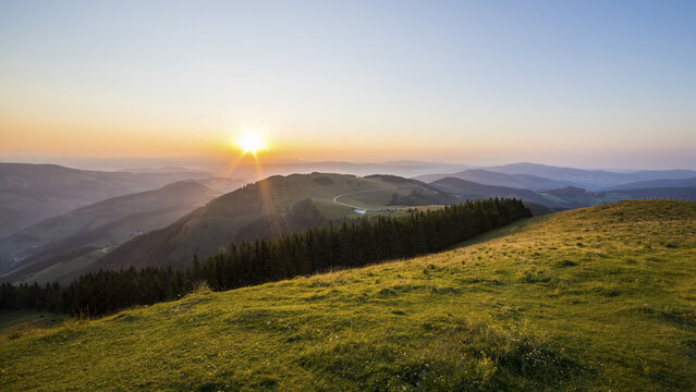 View of the sunset over the Teichalm mountain landscape with rolling grassy hills, a line of pine trees, and distant peaks in Plankogel, Styria, Austria.