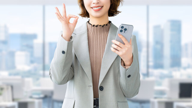 スマートフォンを持ってOKポーズをする笑顔の女性・ビジネスウーマン