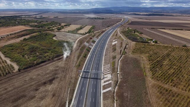 An empty, multi-lane highway cuts through vast agricultural fields under a cloudy sky, suggesting travel and development.