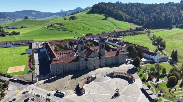 Oblique aerial of Einsiedeln Abbey in Switzerland, showing twin towers, red tiled roofs and a spacious forecourt bordered by gardens and green hills. Scenic religious heritage for tourism and travel.