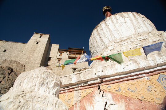 View of a traditional white stupa adorned with colorful prayer flags in front of the historic Leh Palace under a clear blue sky in Leh, Ladakh, India.