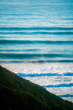 View of rolling ocean waves crashing against the grassy coastal cliffs in Newquay, England, United Kingdom.