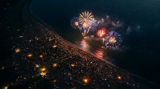 Aerial view of a large crowd gathered on a beach at night watching a spectacular fireworks display over the ocean