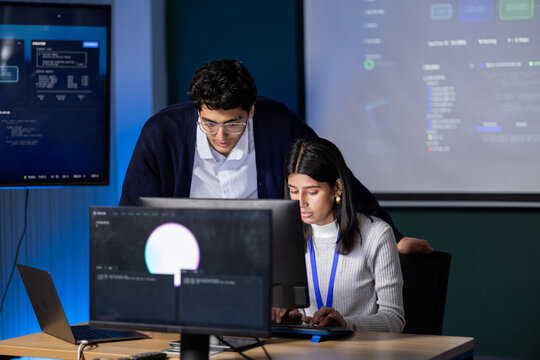 Computer Programmer consults seated developer across multiple monitors, bringing software discussion into shared office work, connecting cooperative computer work with professional activity.