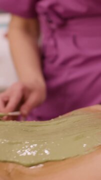 Vertical video. Body care. Anti-aging treatment. Unrecognizable beautician hand applying alginate mask mixture on woman belly in beauty professional clinic.