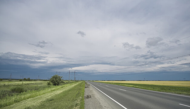 An asphalt highway amidst green fields under a cloudy sky, with power lines running across the fields&mdash;a summer landscape featuring a major transport route