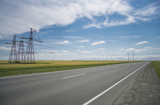 An asphalt highway amidst green fields under a cloudy sky, with power lines running across the fields&mdash;a summer landscape featuring a major transport route
