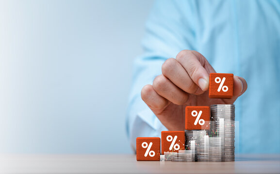Hand stacking percentage symbols on rising coin piles with financial charts for increasing interest rates, financial investment returns growth from banking and economic trends changes.