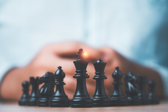 Black chess king and pieces with hands of a strategist in the background, symbolizing business leadership strategy planning, competition and successful management.