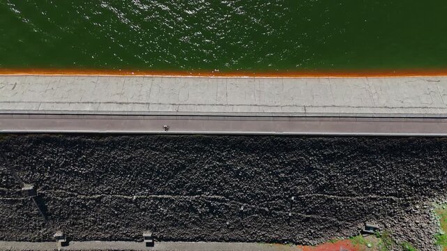 Cyclist rides along a narrow dam road separating green reservoir water from dark riprap, aerial top-down