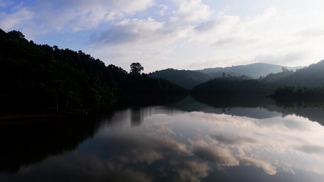 Visitors enjoy the view of Da Lay Irrigation Reservoir in Lam Dong, Vietnam. The water reflects mountains and trees as the sun rises. People walk and take pictures near the shore.