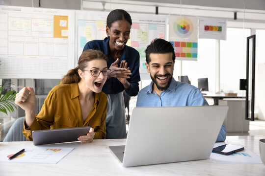 Three cheerful coworkers celebrate successful project completion or breakthrough while looking at laptop screen in modern office, feeling excitement, enjoying good work result, sales growth, promotion