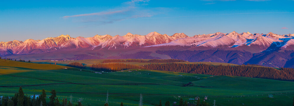 Aerial view of golden sunset light illuminating snow-capped mountain peaks and green forests in Kalajun Grassland, Xinjiang, China.