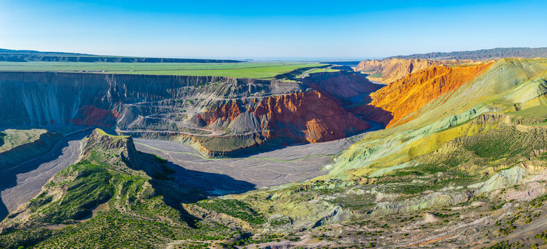 Aerial view of colorful mineral-layered rock cliffs and a winding riverbed under a sunny sky in Anjihai Grand Canyon, Xinjiang, China
