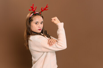 Confident Asian woman wearing reindeer headband flexing arm muscle showing strong power gesture on brown studio background during Christmas.  © Ryan