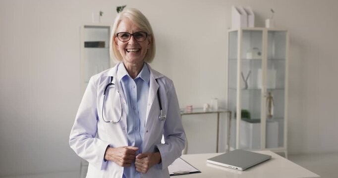 Middle aged female doctor in lab coat standing in personal clinic office looking at camera with confident smile representing professional authority trusted medical expertise premium healthcare service