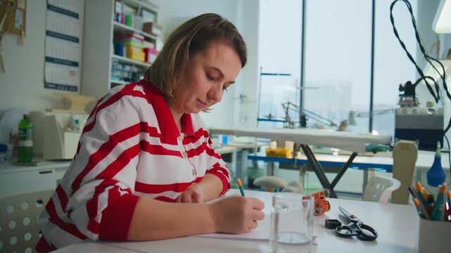 Woman in red striped shirt focuses on drawing at a bright desk. She leans forward, concentrating intently on her work. Tools and glass cup sit near her workspace