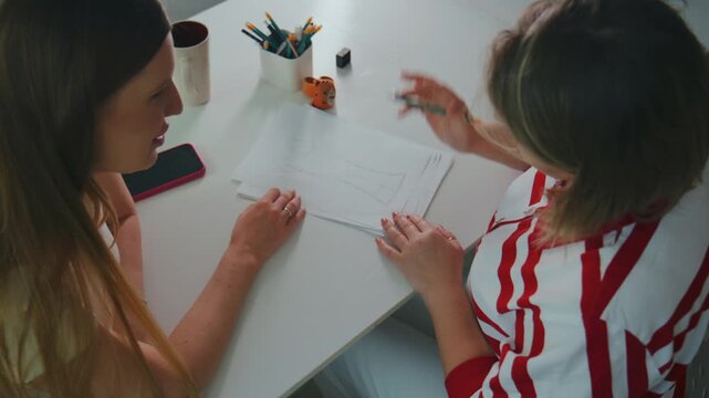Two young women sit at a white table, focused on drawing. One holds a pencil, sketching on paper. The other watches, hands resting near the artwork. Pens and pencils sit in a cup nearby