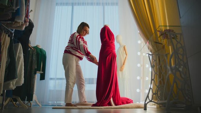 Woman adjusts red wedding dress on mannequin. Soft lighting highlights fabric texture. Behind her, clothes rack stands tall. Yellow curtains frame scene warmly. Ideal for wedding planning, fashion