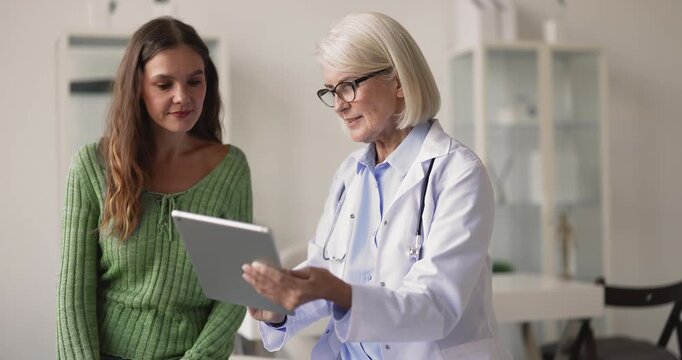 Middle aged female physician discussing diagnostic examination results on digital tablet screen with woman patient explaining medical data in healthcare mobile app on consultation in modern clinic